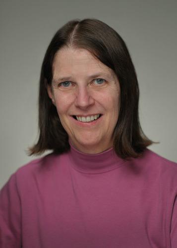 Head-and-shoulders portrait of a person with shoulder-length dark hair wearing a pink high-neck top against a plain gray background.