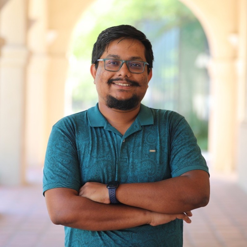 Person with short dark hair, glasses, and facial hair wearing a teal patterned polo shirt and smartwatch, standing with arms crossed in a softly lit arched walkway.