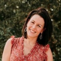 Person with shoulder-length dark hair wearing a sleeveless red patterned top and earrings stands outdoors with greenery in the background.