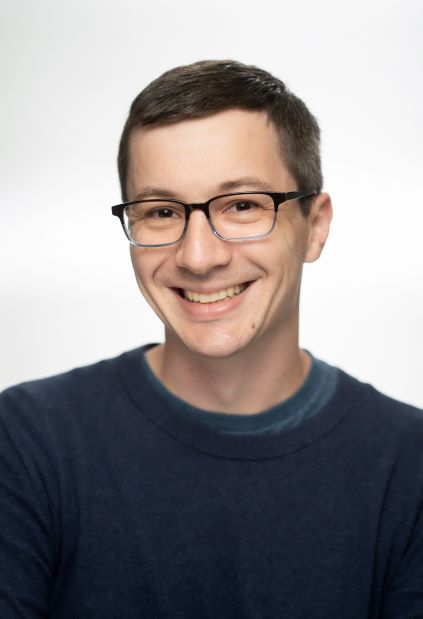 Head-and-shoulders portrait of a person wearing glasses and a dark blue sweater against a light gray background.