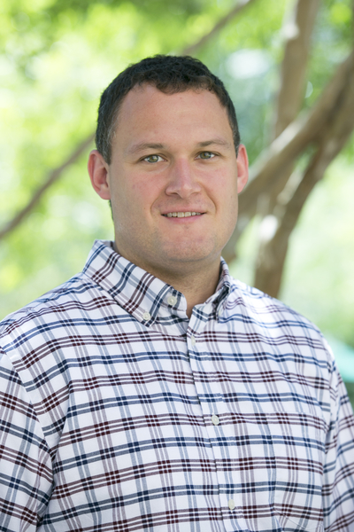 Head-and-shoulders portrait of a person wearing a white plaid button-up shirt, standing outdoors in front of green foliage and tree trunks.