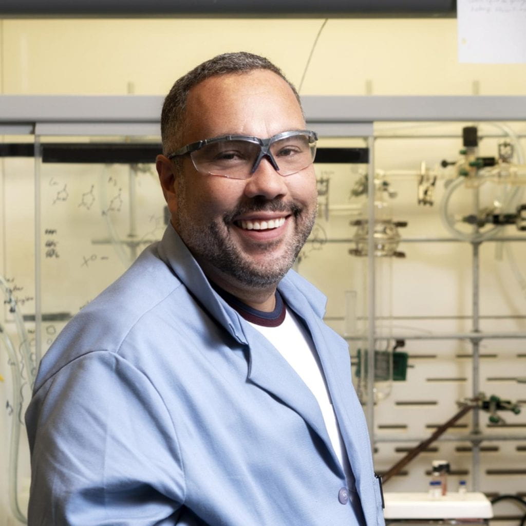 Person wearing safety glasses and a light blue lab coat stands in a chemistry laboratory, with glassware, equipment, and chemical structures visible in the background.
