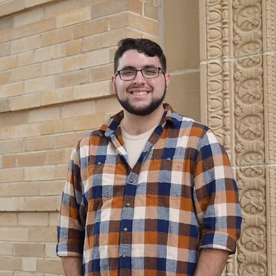 Person wearing glasses and an orange, blue, and white plaid button-up shirt stands in front of a tan brick wall beside a carved stone column.
