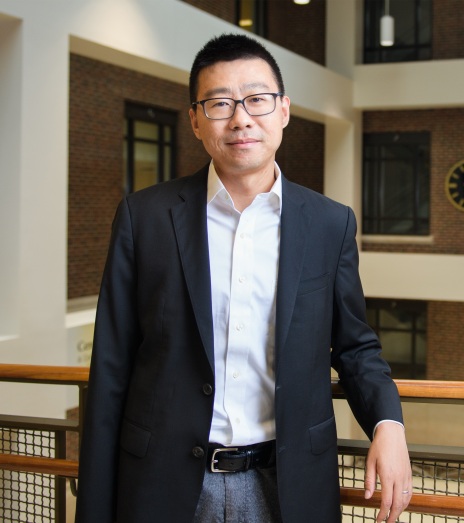 Person wearing glasses, a dark blazer, and a white button-up shirt stands indoors by a railing, with brick walls and windows in the background.