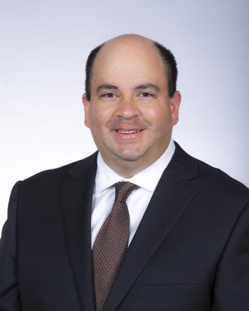 Head-and-shoulders portrait of a person wearing a dark suit, white dress shirt, and patterned tie against a light background.
