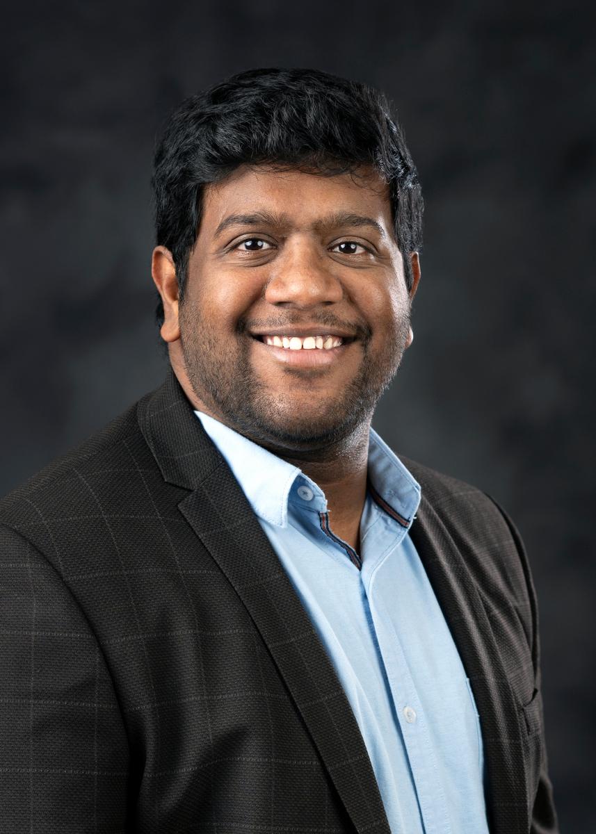 Head-and-shoulders portrait of a person with short dark hair wearing a dark blazer and light blue collared shirt against a dark background.