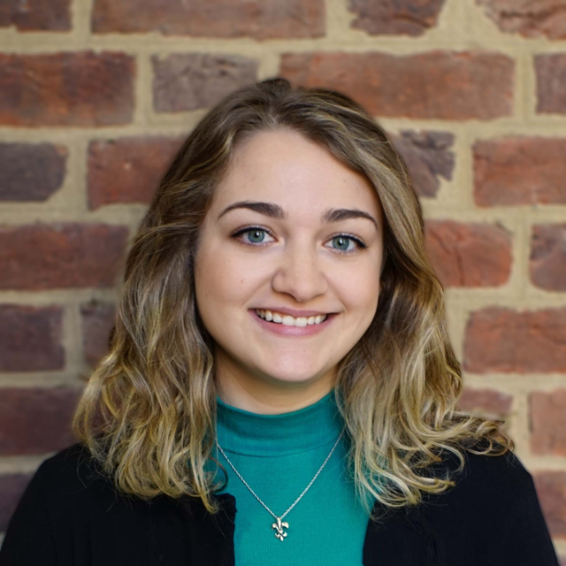 Head-and-shoulders portrait of a person with wavy light brown hair wearing a teal turtleneck, black cardigan, and necklace in front of a brick wall.